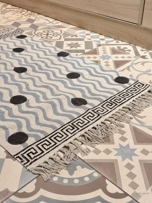 A close-up of a woven area rug with a cream base, light blue wavy stripes, black polka dots, and a black Greek key border with fringe, laid on a patterned tile floor.