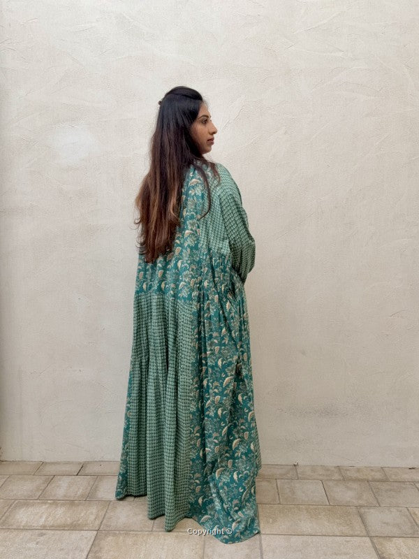 Woman wearing a long green floral dress against a plain wall.