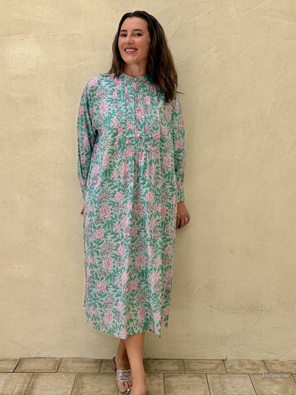 A woman wearing a long-sleeve midi dress with a green and pink floral pattern, standing against a textured beige stucco wall.