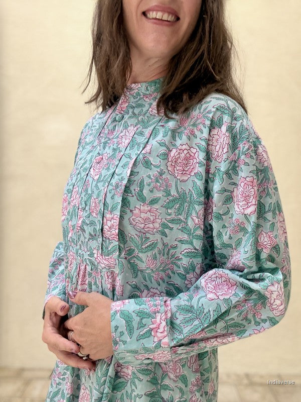 A woman wearing a long-sleeve high-neck tunic dress with a pink and green floral block print on a light teal background, standing against a neutral beige wall.