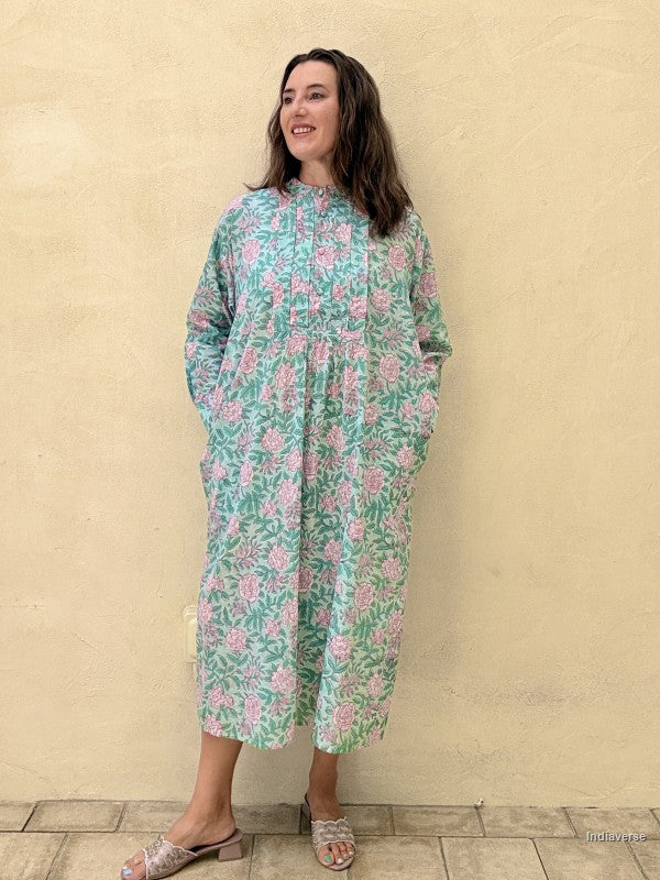 A woman wearing a long-sleeve, high-neck midi dress with a green and pink floral pattern, standing against a textured beige stucco wall.