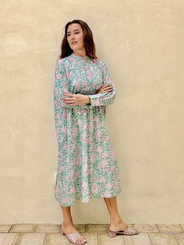 A woman modeling a long-sleeve midi dress with a green, pink, and white floral pattern, standing against a textured beige stucco wall.