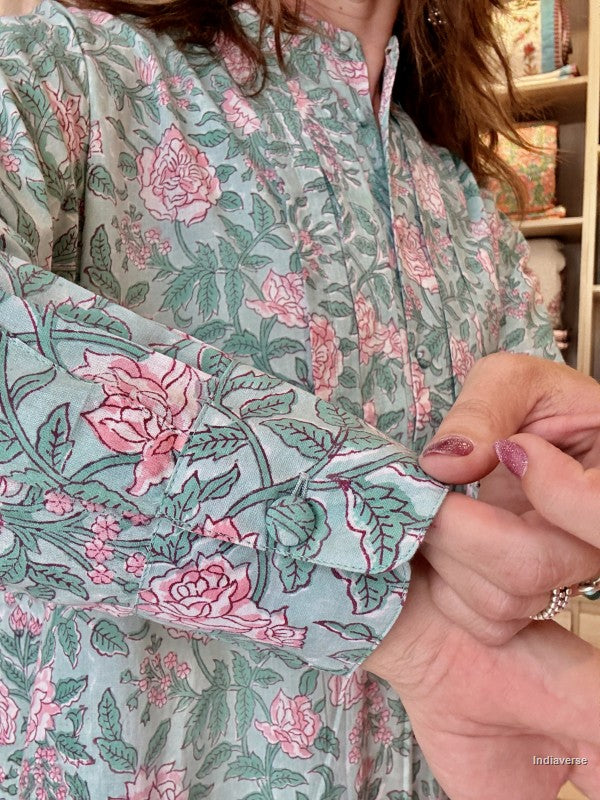A close-up of a person wearing a long-sleeve button-up shirt with a sage green base and a pink and green floral block print. The person's hands are shown adjusting the cuff of the shirt.