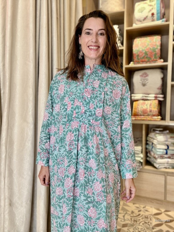 A woman smiling while wearing a long-sleeved, high-neck dress with a green base and pink floral block print, standing in front of a beige curtain and a wooden shelf filled with folded textiles.