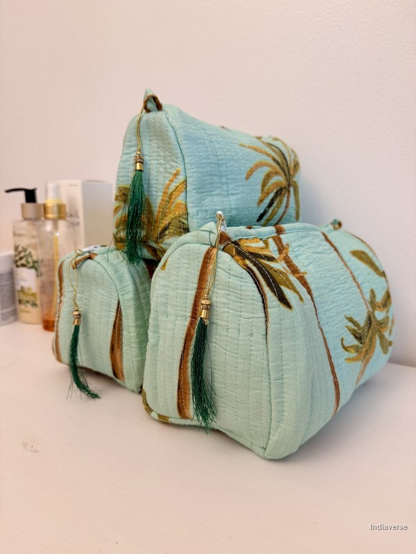 Three light blue fabric cosmetic pouches with gold palm tree embroidery and green tassels, arranged on a white surface with toiletry bottles in the background | Turquoise Palm