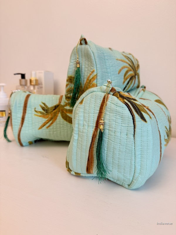 Three light aqua quilted cosmetic pouches with a gold palm tree print and green tassel zipper pulls, arranged on a white surface against a pale pink background | Turquoise Palm