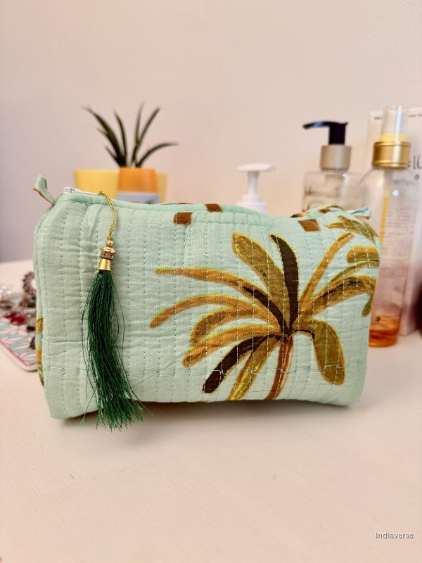 A light mint green quilted cosmetic pouch with a large gold and brown palm leaf print, featuring a dark green tassel zipper pull, set on a tabletop with skincare bottles in the background | Turquoise Palm