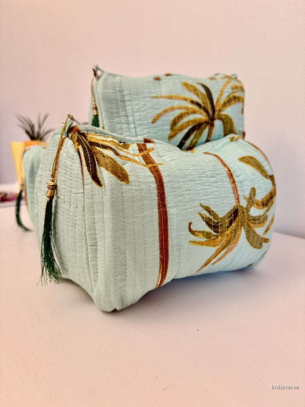 A light blue quilted fabric pouch with embroidered palm tree motifs in gold and brown, featuring a dark green tassel zipper pull, set against a soft pink background | Turquoise Palm