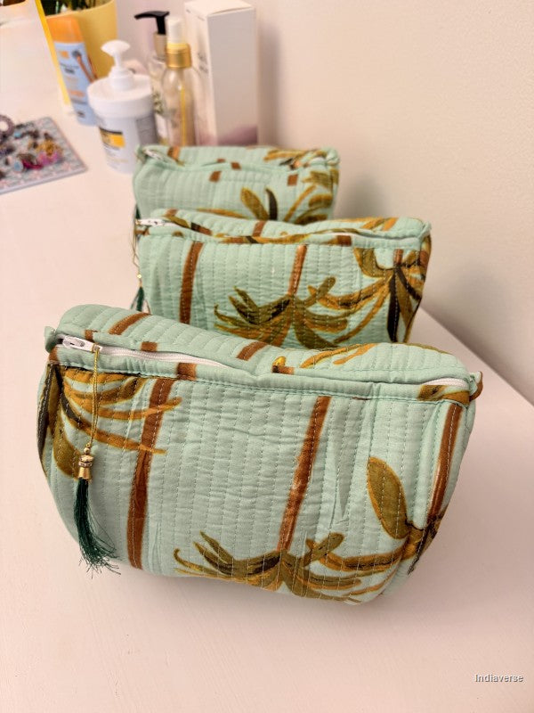 Three quilted cosmetic pouches with a bamboo leaf pattern in sage green, brown, and gold, arranged on a light pink surface with toiletries in the background | Turquoise Palm
