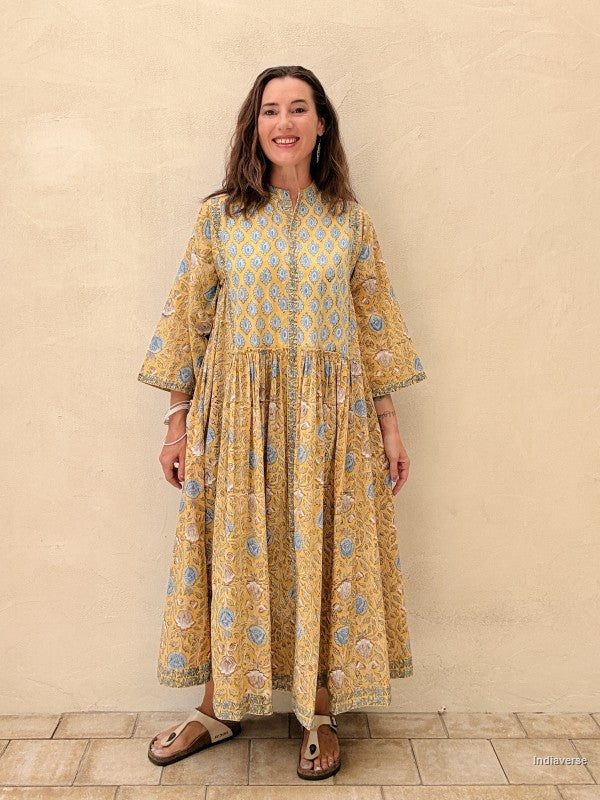 A woman modeling a long-sleeved, tiered maxi dress with a yellow and blue geometric pattern, standing against a textured beige stucco wall.