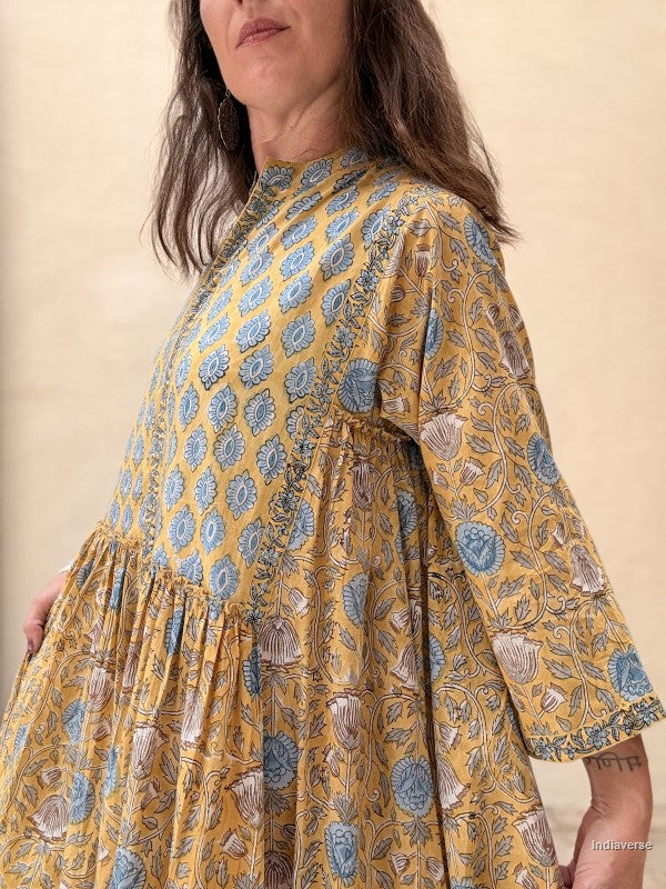A woman wearing a mustard yellow dress with a blue and gray floral block print, featuring a high neckline and elbow-length sleeves, set against a plain beige background.