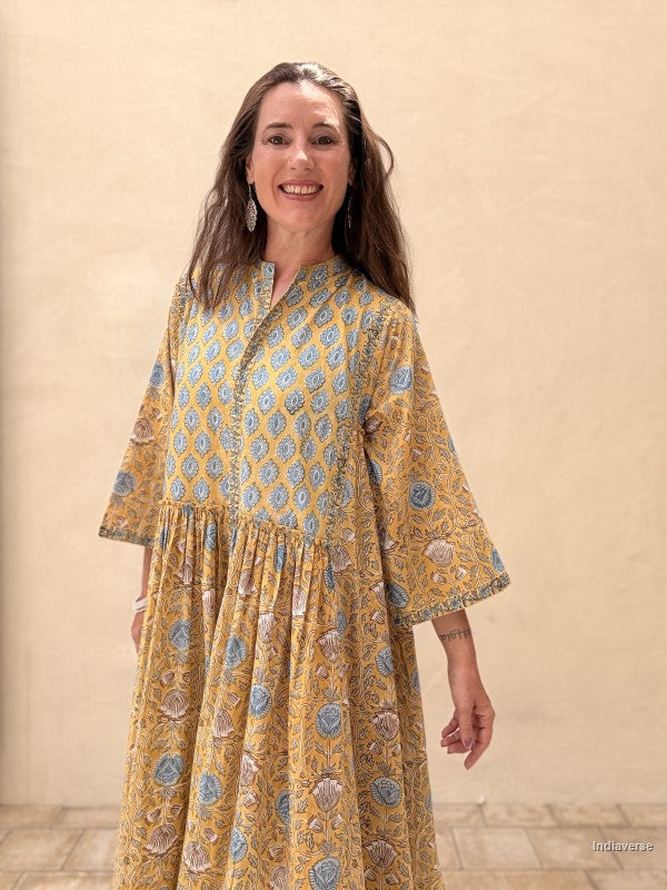 A woman wearing a mustard yellow kaftan dress with blue and beige floral block prints, standing against a neutral beige stucco wall.