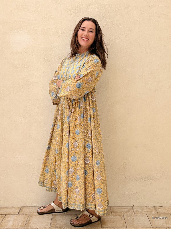 A woman wearing a long-sleeved, high-neck maxi dress with a yellow and blue paisley pattern, standing against a beige stucco wall.