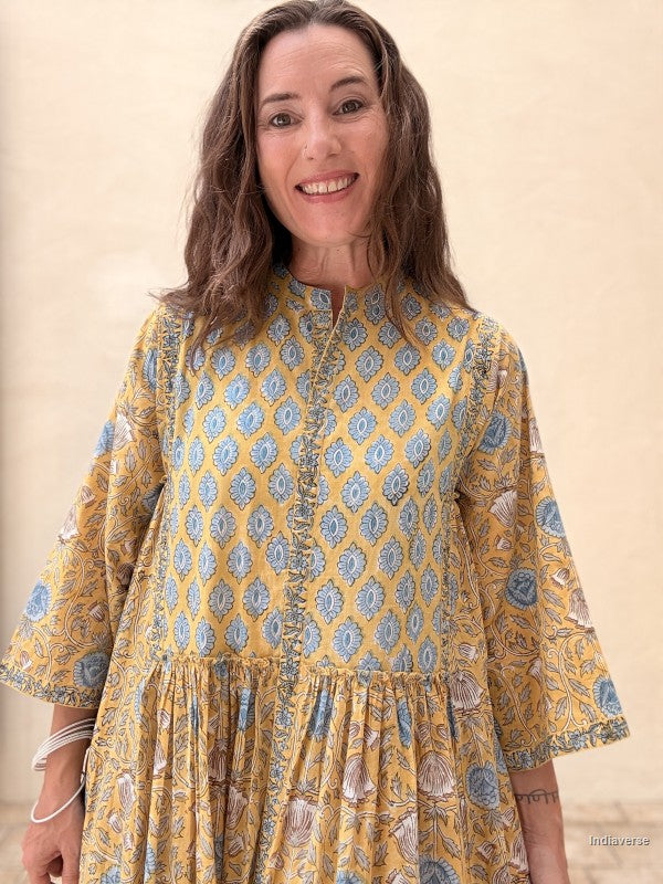 A woman wearing a yellow and blue patterned cotton tunic dress with three-quarter sleeves, standing against a neutral beige background.