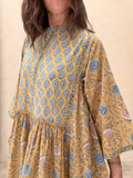 A woman wearing a mustard yellow tiered dress with a blue and gray block print pattern, shown from the neck down against a neutral beige background.