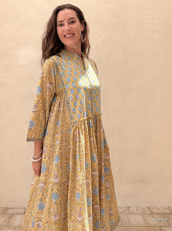 A woman wearing a yellow and blue patterned dress stands against a beige stucco wall, smiling. The dress has a loose, flowing silhouette with three-quarter sleeves and an allover floral block print.