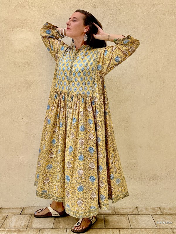 A woman wearing a long-sleeved, tiered kaftan dress with a blue and gold geometric pattern, standing against a textured beige stucco wall.