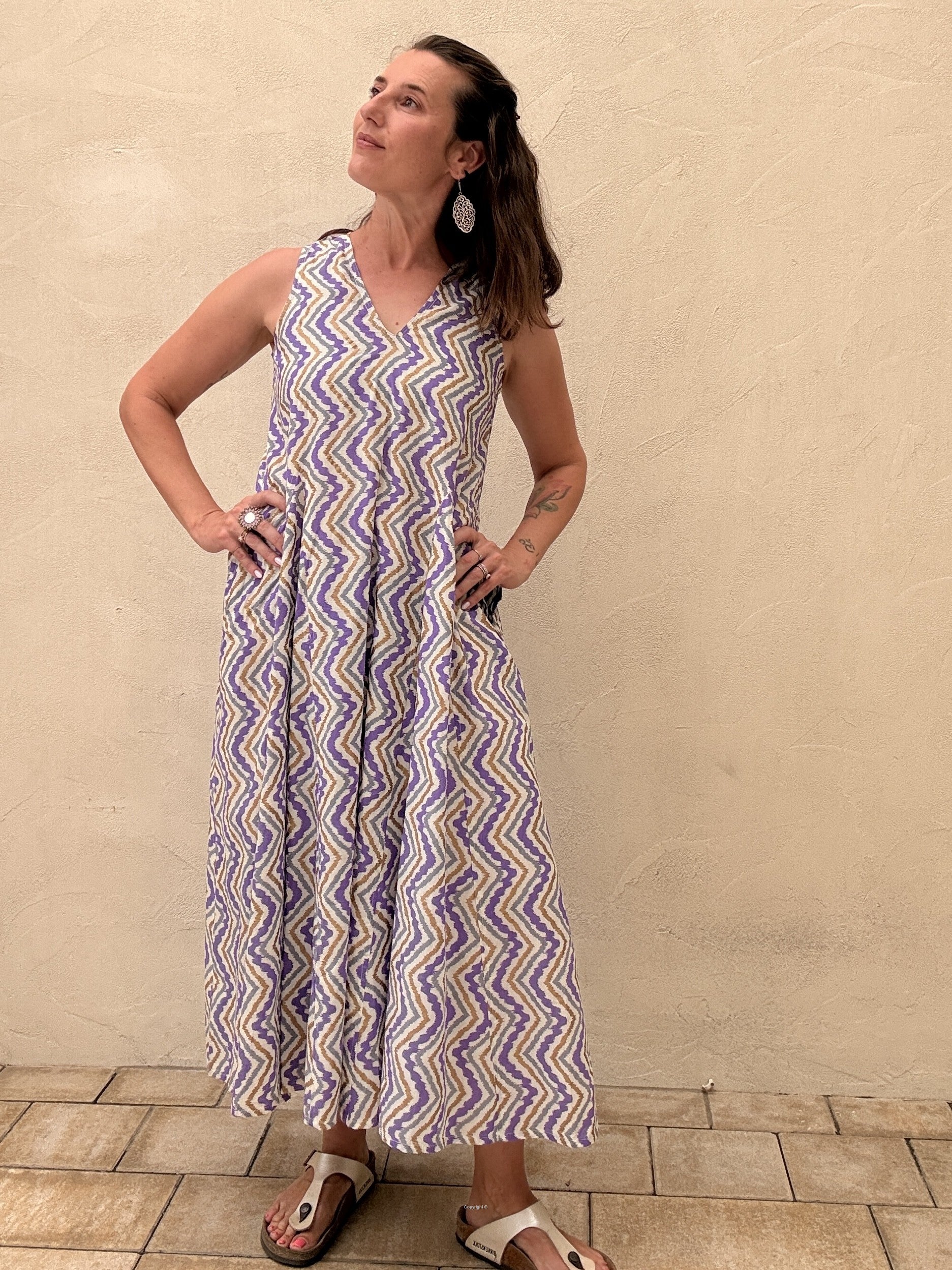Woman wearing a sleeveless, long-patterned dress against a beige wall.
