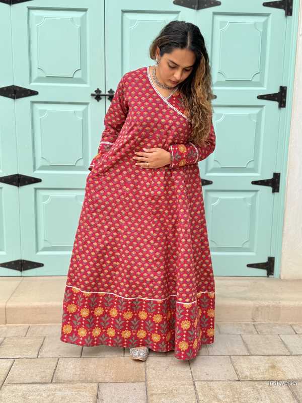 Woman wearing a full length ethnic festive wrap dress with silver border around the neckline and arms and floral trim border in front of light blue colored door