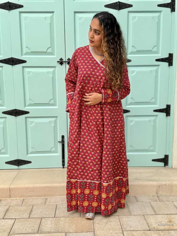 Woman wearing a full length ethnic festive wrap dress with silver border around the neckline and arms and floral trim border in front of light blue colored door