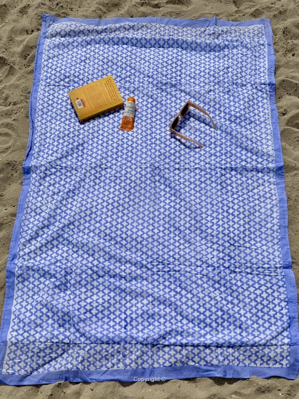 Blue patterned towel on sand with a yellow sponge, orange bottle, and brown tool.