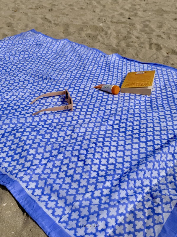 Blue and white patterned towel on sand with a bottle, clip, and book.