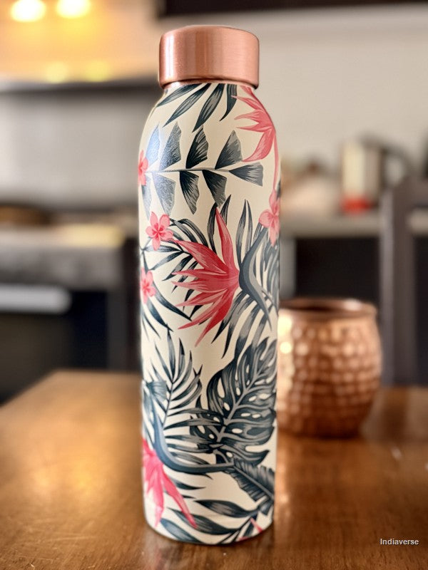 copper water bottle with tropical leaf and birds of paradise design sitting on a brown surface in a kitchen with a copper mug in the background