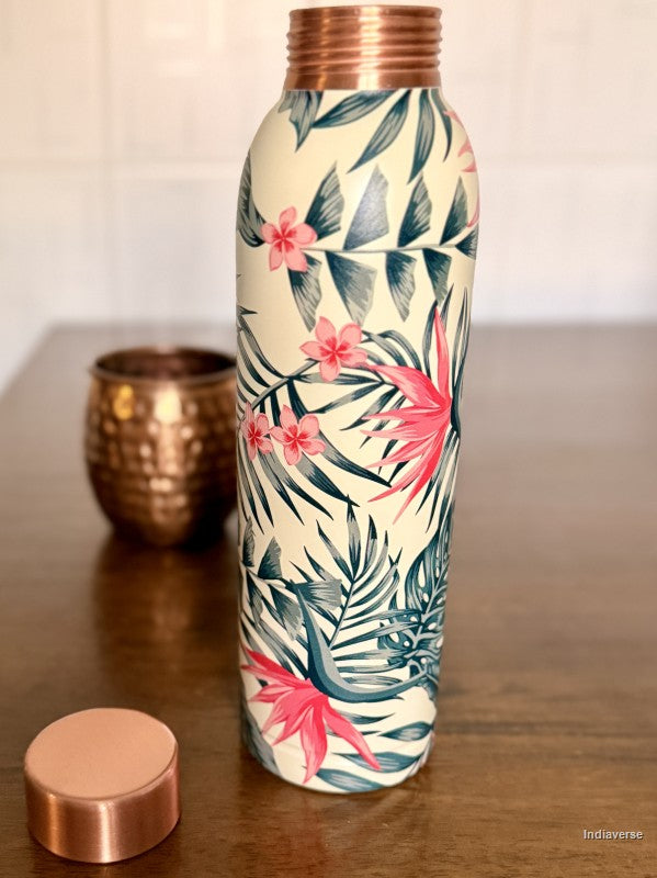 copper water bottle with tropical leaf and birds of paradise design sitting on a brown surface with a copper mug in the background
