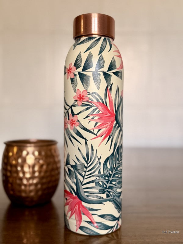 copper water bottle with tropical leaf and birds of paradise design sitting on a brown surface with a copper mug in the background