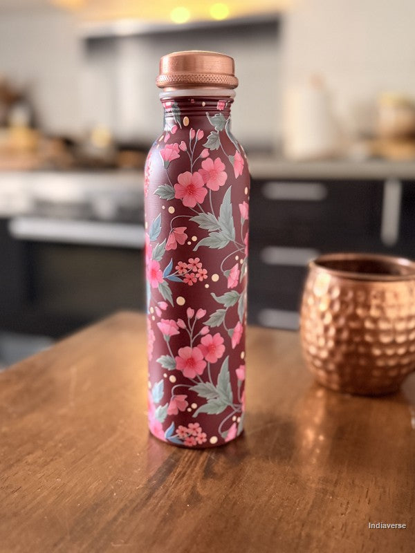 copper water bottle with red and pink floral design and a Bordeaux color background sitting on a kitchen table with a copper cup in the background
