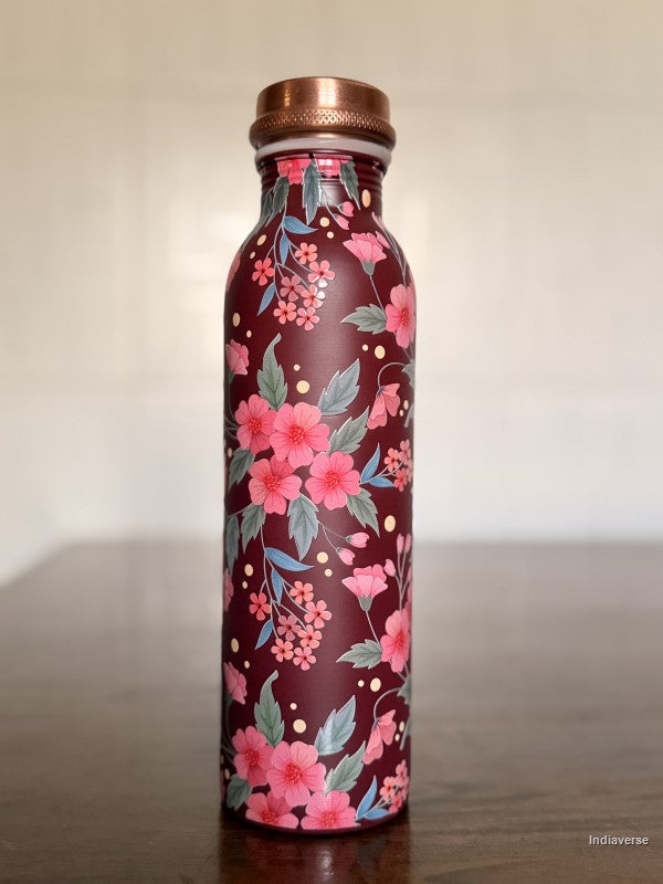 copper water bottle with red and pink floral design and a Bordeaux color background sitting on a brown table