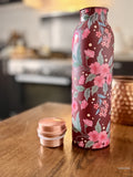 copper water bottle with red and pink floral design and a Bordeaux color background sitting on a kitchen table with a copper cup in the background
