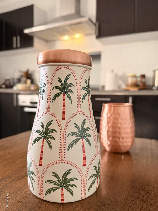 A decorative copper water jar with geometric palm tree design is sitting on a brown table in a kitchen with a copper mug in the background