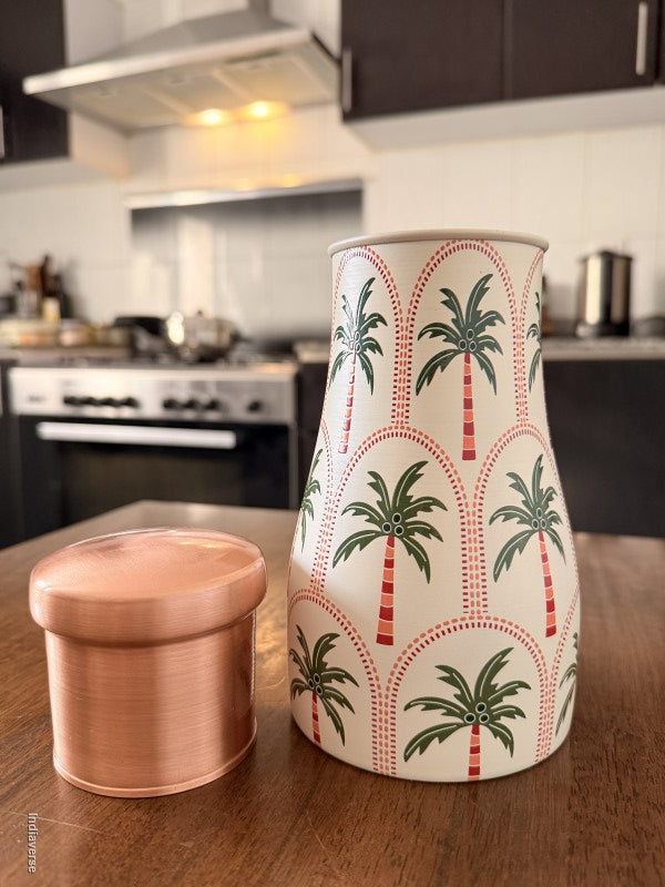 A decorative copper water jar with geometric palm tree design is sitting on a brown table with its copper lid on the side