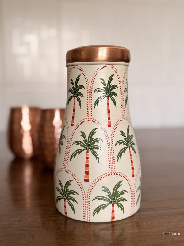 A decorative copper water jar with geometric palm tree design is sitting on a brown surface with two copper mugs in the background