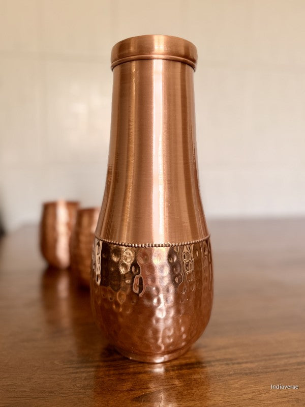 tall pure copper water jar with hammered bottom and smooth copper body sitting on a brown table with two copper cups in the background