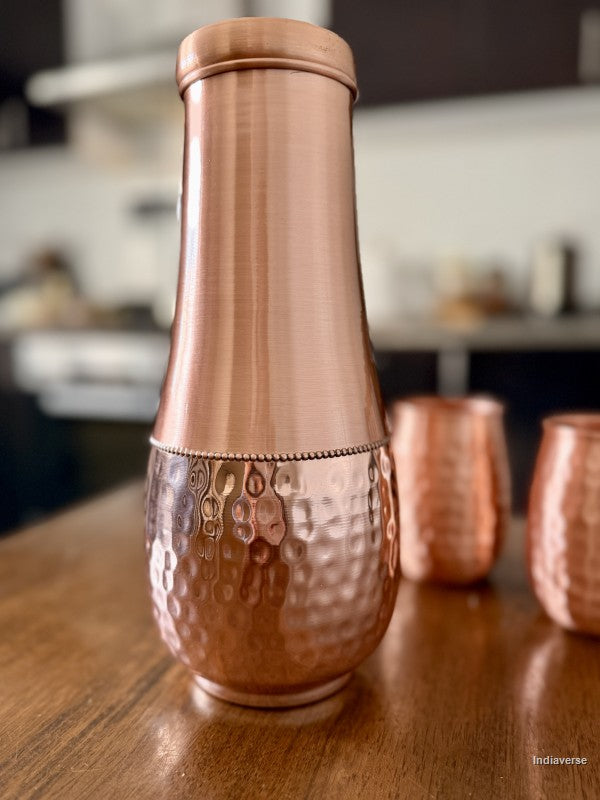 tall pure copper water jar with hammered bottom and smooth copper body sitting on a brown table in a kitchen with two cups in the background
