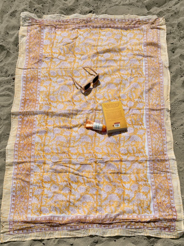 Yellow patterned blanket on sand with sunglasses and a book