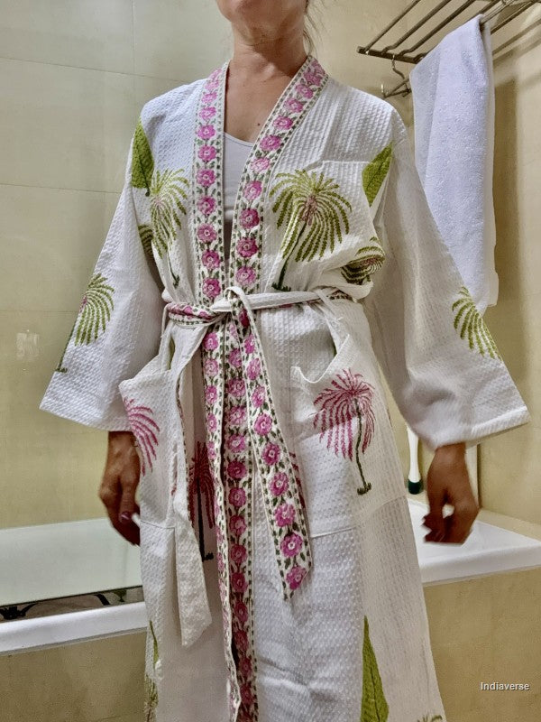 A person wearing a white waffle-knit bathrobe with pink floral trim and green palm tree embroidery, standing in a bathroom with a bathtub and towel visible.
