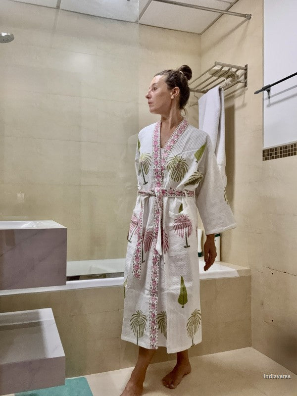 A woman wearing a white cotton bathrobe with pink and green palm tree prints stands in a beige tiled bathroom.