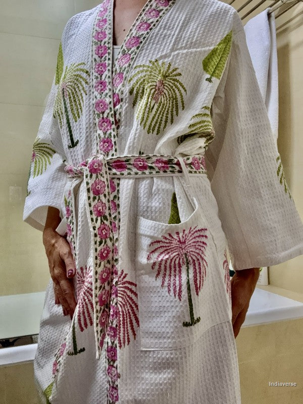 A white waffle-knit robe with pink and green palm tree prints and a matching patterned tie belt, shown on a person in a bathroom setting.