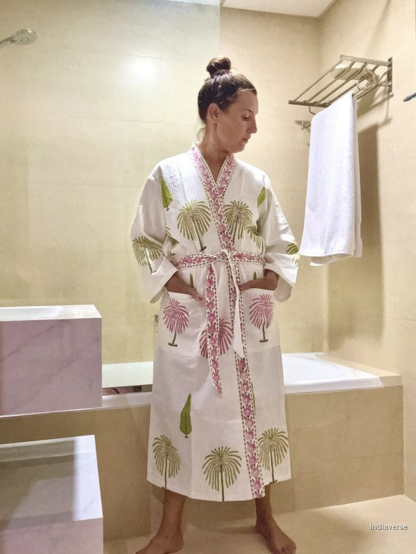A woman wearing a white cotton bathrobe with pink and green palm tree prints, standing in a beige tiled bathroom.