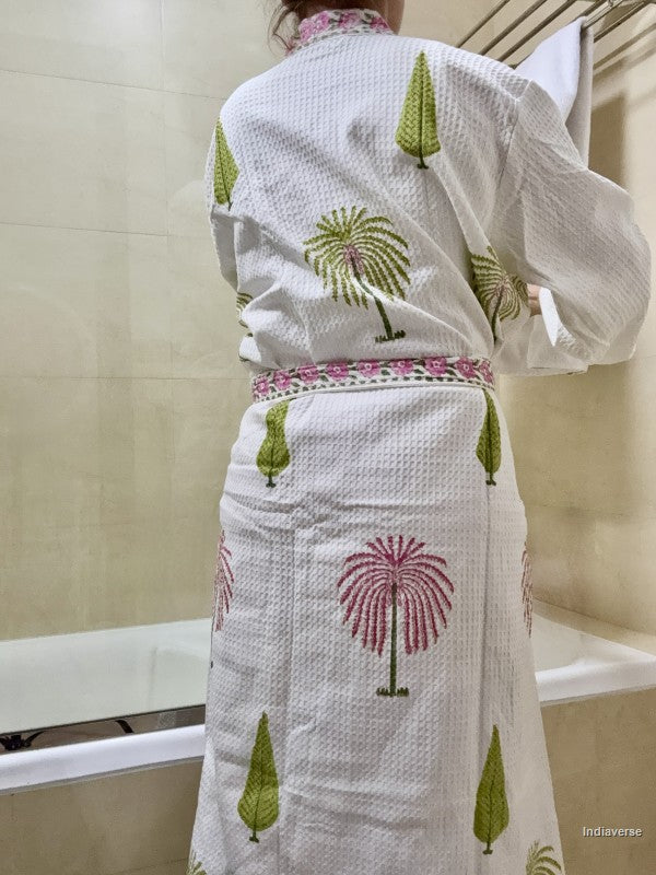 A person viewed from behind wearing a white textured bathrobe with green and pink palm tree prints and a floral embroidered waistband, standing in a bathroom.