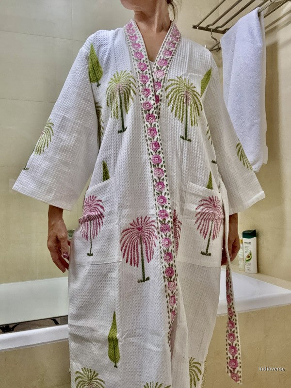 A woman wearing a white waffle-knit robe with pink and green palm tree prints and a floral embroidered border, standing in a bathroom.