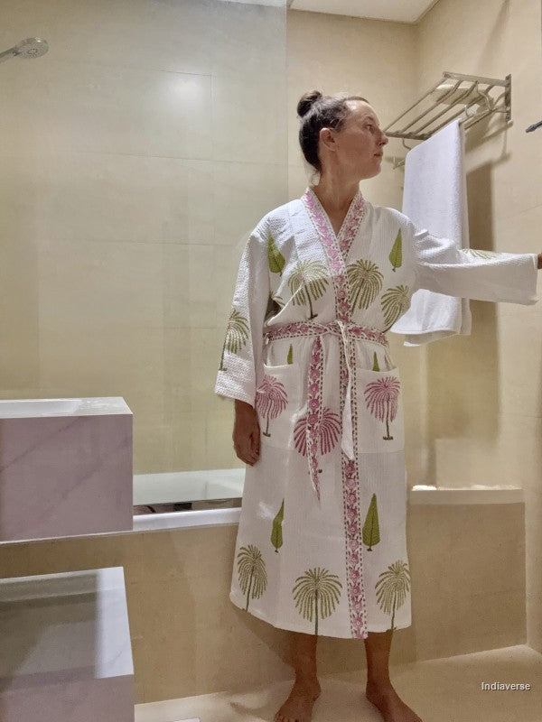 A woman wearing a white cotton bathrobe with pink and green palm tree patterns stands in a beige tiled bathroom next to a bathtub.
