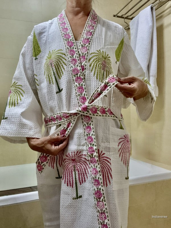 A person wearing a white textured bathrobe with pink floral borders and green palm tree prints, tying the belt at the waist in a bathroom setting.