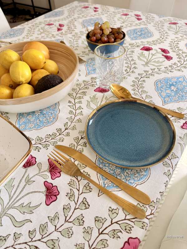 Dining table setting with blue plate, gold cutlery, and bowl of fruit on a floral tablecloth.
