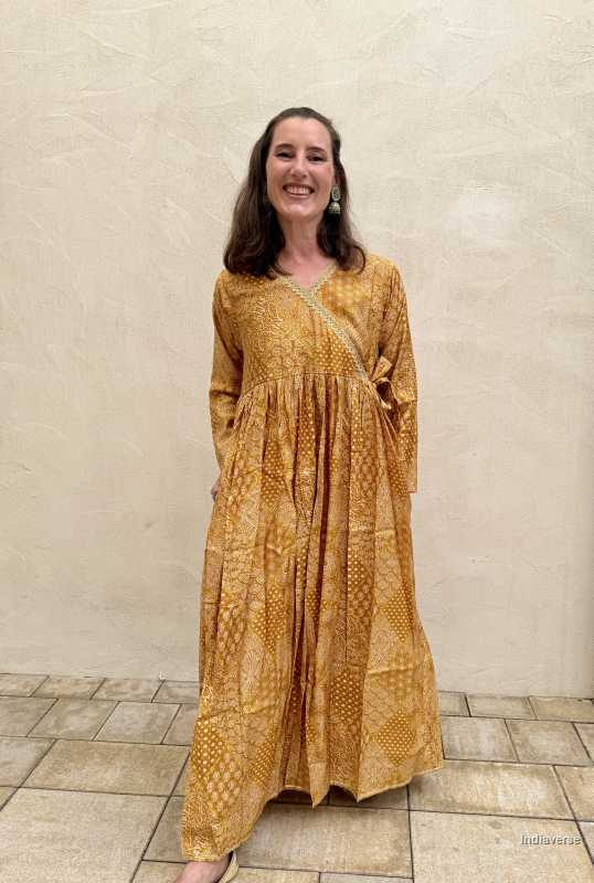 Woman wearing a mustard yellow patterned dress standing against a beige wall.