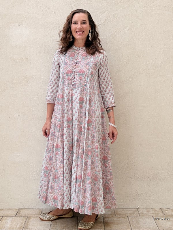 A woman modeling a long-sleeved, high-neck maxi dress with a pink, blue, and white floral block print, standing against a textured beige stucco wall.