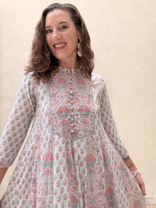 A woman wearing a long-sleeved, high-neck A-line tunic dress with a pink, green, and white block print pattern, standing against a plain beige wall.